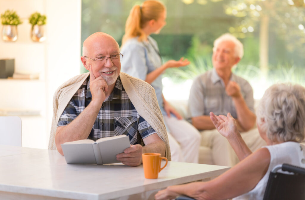 An older adult man sitting at a table holding a book and engaged in conversation with an older adult woman in a wheelchair.