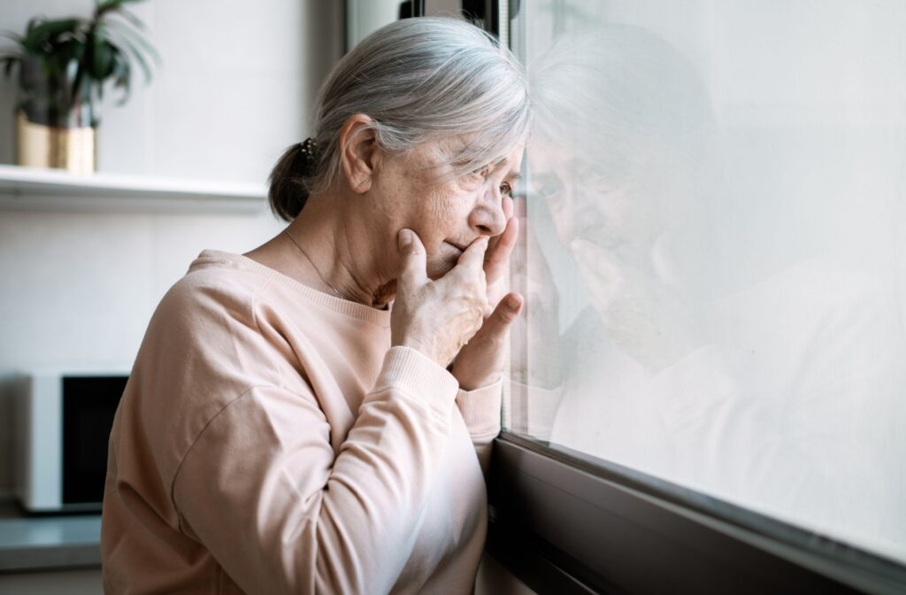 A worried senior hold their hand to their mouth as they gaze out the window, trying to recall an important upcoming event